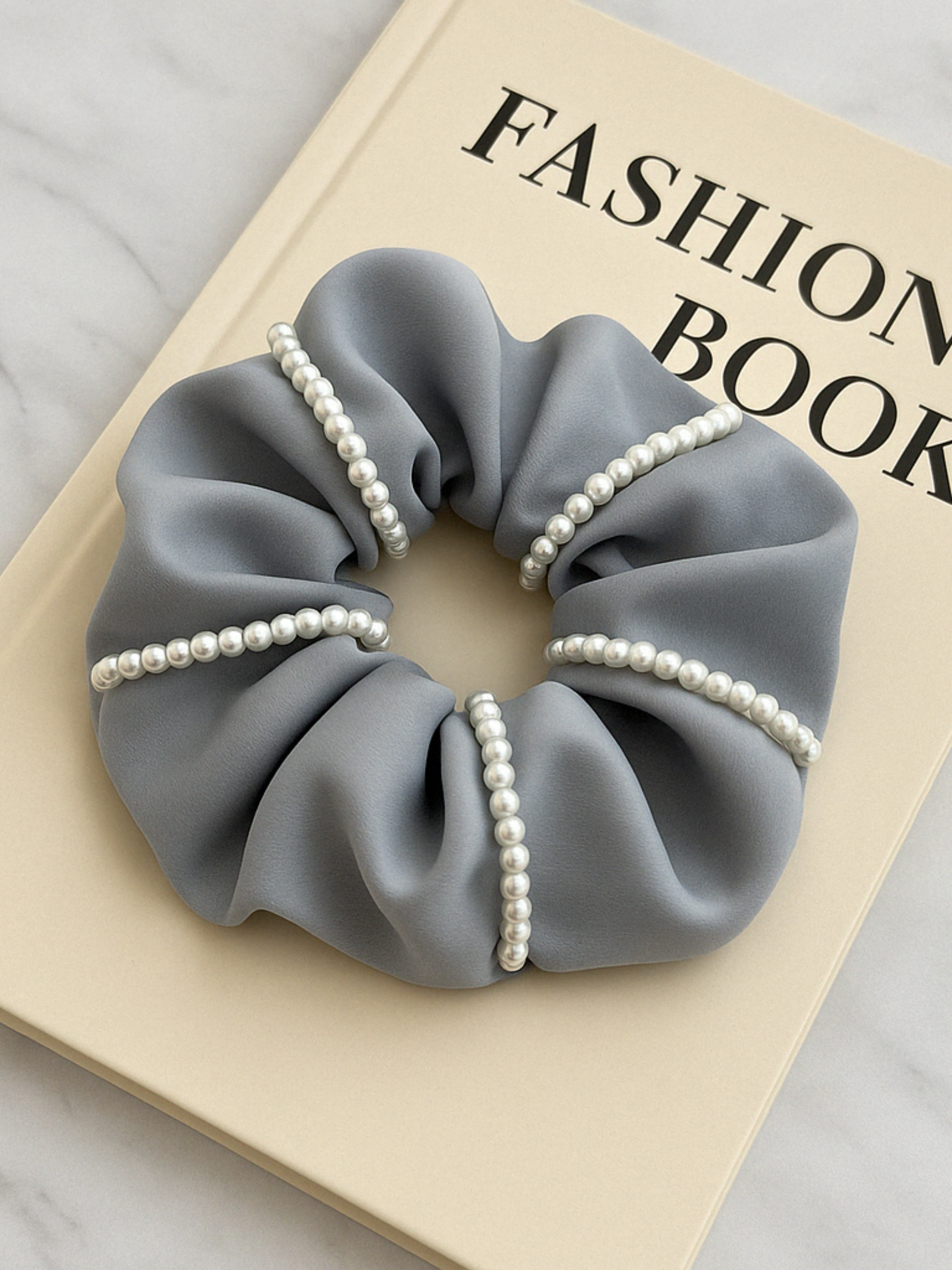 Chouchou en satin gris orné de perles, accessoire élégant pour cheveux, posé sur un livre de mode.